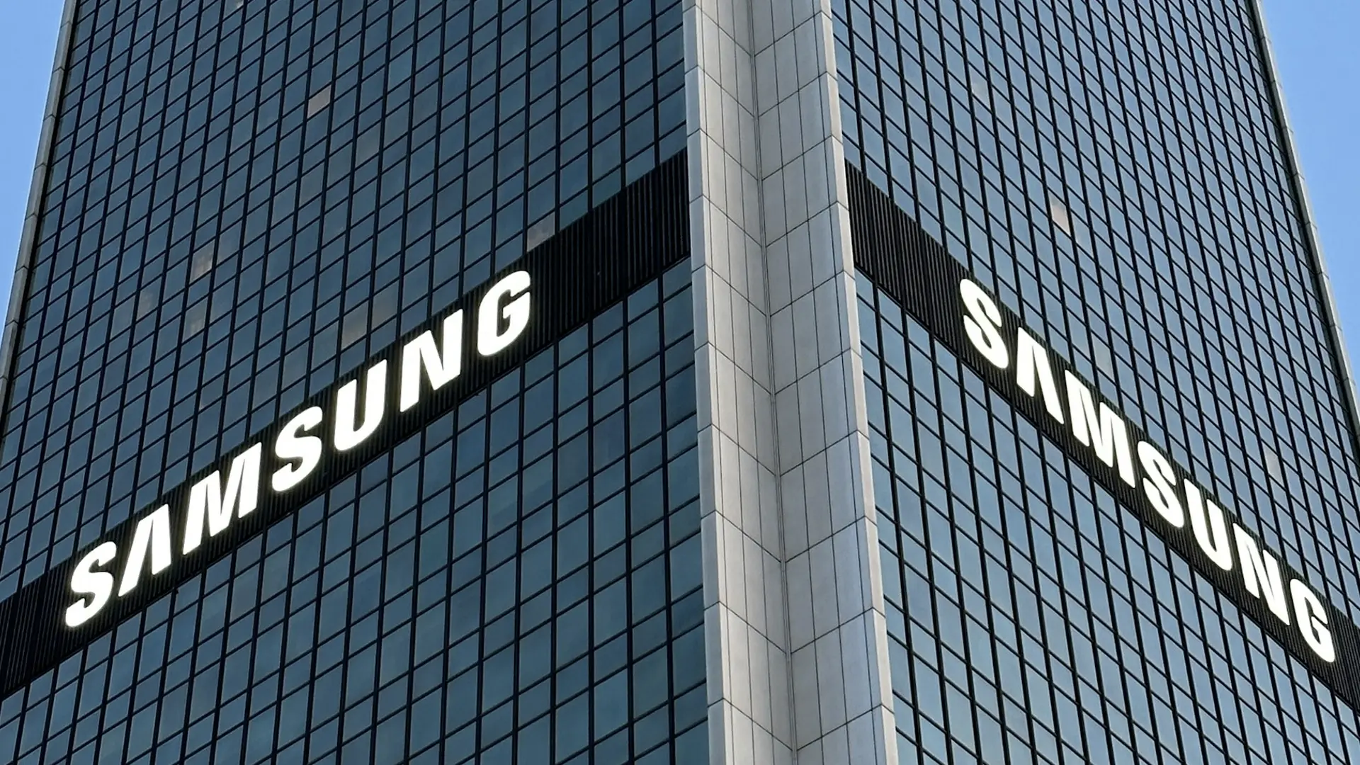 Corner of a glass office tower with large white SAMSUNG logos on two sides seeing the building at an angle.