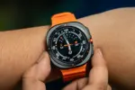 Close-up of an orange-strapped smartwatch on a wrist, displaying a busy analog-digital watch face with orange accents.