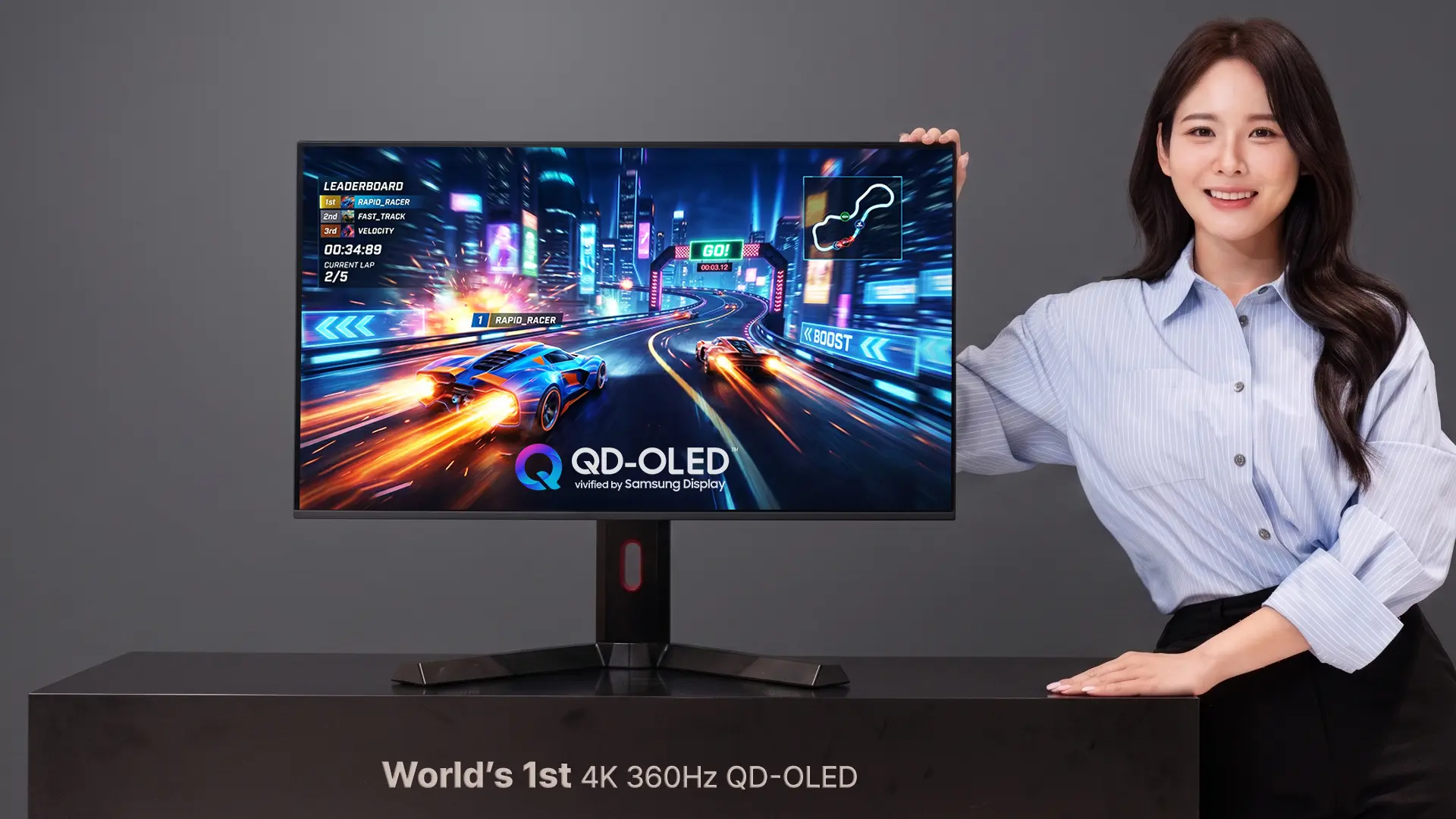 Woman posing with a large gaming monitor showing a neon-race car game on a desk. The display reads QD-OLED and 4K 360Hz markers are visible.