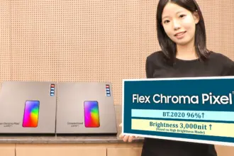 Woman standing behind a counter, holding a large sign that reads 'Flex Chroma Pixel' while two boxed devices sit on the counter beside her.